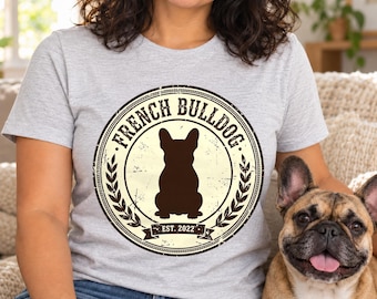 May include: A light gray t-shirt featuring a circular graphic with a dark brown silhouette of a French Bulldog in the center. The text reads FRENCH BULLDOG and EST. 2022, surrounded by a decorative laurel leaf border in a distressed style.