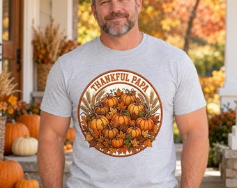 May include: A light gray short-sleeve t-shirt featuring a circular graphic on the chest. The design displays a collection of orange pumpkins, brown autumn leaves, and wheat stalks, with the text Thankful Papa arched across the top.