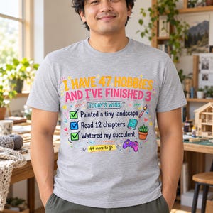 May include: A heather gray short-sleeve t-shirt featuring the text I HAVE 47 HOBBIES AND I'VE FINISHED 3. Below, a checklist reads TODAY'S WINS with three green and blue checkmarks next to painted a tiny landscape, read 12 chapters, and watered my succulent.
