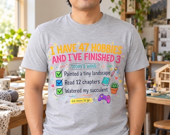 May include: A heather gray short-sleeve t-shirt featuring the text I HAVE 47 HOBBIES AND I'VE FINISHED 3. Below, a checklist reads TODAY'S WINS with three green and blue checkmarks next to painted a tiny landscape, read 12 chapters, and watered my succulent.