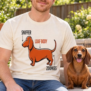 May include: A cream-colored short-sleeve t-shirt featuring a graphic of a brown dachshund. The design includes black text pointing to the dog's nose labeled SNIFFER, orange text pointing to the back labeled LOAF BODY, and black text pointing to the tail labeled ZOOMIES.