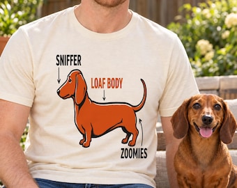 May include: A cream-colored short-sleeve t-shirt featuring a graphic of a brown dachshund. The design includes black text pointing to the dog's nose labeled SNIFFER, orange text pointing to the back labeled LOAF BODY, and black text pointing to the tail labeled ZOOMIES.