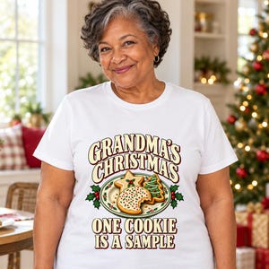 May include: A white short-sleeve t-shirt featuring a graphic of a plate of Christmas cookies with holly leaves. The text reads: "GRANDMA'S CHRISTMAS ONE COOKIE IS A SAMPLE" in a bold, dark red and cream-colored font style.