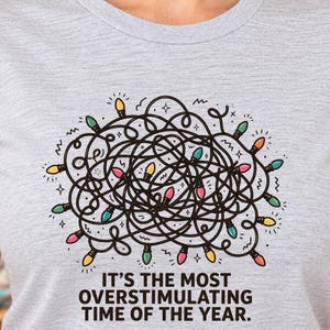 May include: A heather gray t-shirt featuring a graphic of a tangled black line drawing of Christmas lights with colorful bulbs in yellow, pink, green, and blue. Below the graphic, the text reads: IT'S THE MOST OVERSTIMULATING TIME OF THE YEAR.