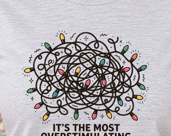 May include: A heather gray t-shirt featuring a graphic of a tangled black line drawing of Christmas lights with colorful bulbs in yellow, pink, green, and blue. Below the graphic, the text reads: IT'S THE MOST OVERSTIMULATING TIME OF THE YEAR.