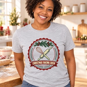 May include: A light gray short-sleeve t-shirt featuring a circular graphic with a red scalloped border. The design includes a whisk and rolling pin, pine branches with pinecones, and the text CHRISTMAS BAKING CREW on a red banner.