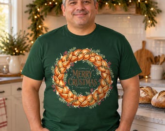 May include: A dark green short-sleeve t-shirt featuring a graphic of a braided bread wreath decorated with pine needles and red berries. The centre of the wreath contains the text Merry Crustmas in a decorative gold-coloured font.