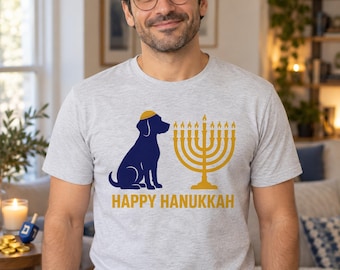 May include: A light gray t-shirt featuring a dark blue silhouette of a dog wearing a yellow yarmulke, positioned next to a yellow menorah with lit candles. Below the graphic, the text reads HAPPY HANUKKAH in bold yellow capital letters.