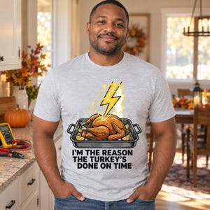 May include: A light gray crew neck t-shirt featuring a graphic of a roasted turkey in a pan struck by a yellow lightning bolt, with the text "I'M THE REASON THE TURKEY'S DONE ON TIME" printed in black capital letters below the illustration.