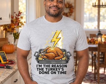 Puede incluir: Una camiseta gris claro de cuello redondo con un gráfico de un pavo asado en una bandeja golpeado por un rayo amarillo, con el texto "I'M THE REASON THE TURKEY'S DONE ON TIME" impreso en letras mayúsculas negras bajo la ilustración.