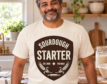 May include: A cream-coloured short-sleeve t-shirt featuring a dark brown shield-shaped graphic on the chest. The text reads SOURDOUGH STARTER, EST. 2020, and FED DAILY, accompanied by illustrations of wheat stalks and a rolling pin.