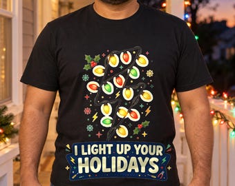 Pode incluir: Uma t-shirt preta de manga curta com um gráfico em forma de relâmpago formado por luzes de Natal coloridas, rodeado por azevinho e flocos de neve, com o texto I LIGHT UP YOUR HOLIDAYS impresso em letras amarelas.
