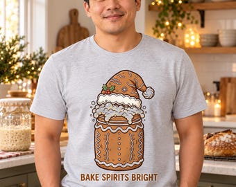 May include: A light grey short-sleeved t-shirt featuring a graphic of a gingerbread-style jar wearing a Santa hat with white foam spilling over the top. The text BAKE SPIRITS BRIGHT is printed in brown letters below the jar illustration.