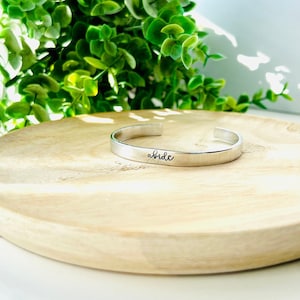 May include: A silver bracelet with the word "abide" engraved on it. The bracelet is sitting on a wooden plate.