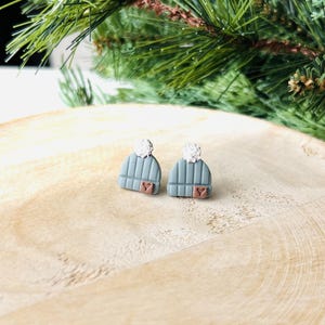 May include: A pair of blue beanie earrings with white pom-poms and a small heart detail. The earrings are set on a wooden surface with green pine branches in the background. The earrings are a winter-themed accessory.