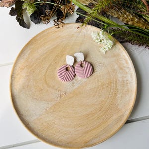 Puede incluir: Un par de pendientes de arcilla geométricos de color rosa y blanco. Los pendientes tienen forma de círculo con un centro blanco y líneas geométricas rosas.