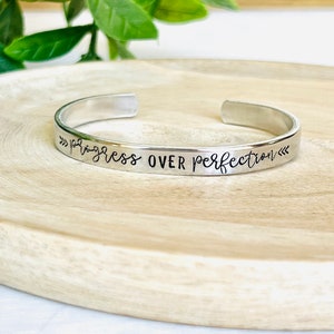 May include: Silver bangle bracelet with the inscription "Progress over perfection" on a wooden surface.
