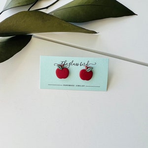 May include: A pair of red apple earrings with green leaves. The earrings are handmade and are displayed on a white card with the text "the glass bird" and "handmade jewelry".
