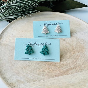 May include: Two pairs of handmade Christmas tree earrings on light blue cards. One pair is a light beige color, and the other is a teal green. The cards read "the glass bird" and "HANDMADE JEWELRY". The earrings are on a wooden tray.