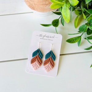 May include: A pair of dangle earrings with a layered leaf design. The leaves are made of polymer clay and are in shades of teal, brown, and pink. The earrings are hanging from gold-toned hooks. The earrings are displayed on a white card with the text "the glass bird handmade jewelry".