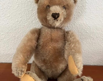 Puede incluir: Un oso de peluche de color marrón claro con pelaje suave y texturizado, nariz bordada en marrón oscuro y parches de tela beige claro en las plantas de los pies. El oso tiene orejas redondeadas, ojos oscuros y una postura sentada.