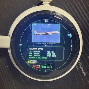 May include: A circular white device with a digital screen displaying a radar-style interface. The screen shows an airplane graphic, flight data for ETD87A B789, altitude of 36000 ft, speed of 541 kt, and the route from Lisbon to Abu Dhabi.