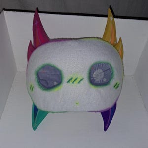May include: A plush toy with a white fuzzy face, two purple and pink horns on top, and two teal and purple horns on the bottom. The face features large purple mesh eyes with white star accents, green stitched blush marks, and a small green dot mouth.