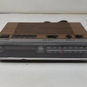 May include: A General Electric clock radio with a wood grain top panel, a black front display, and a horizontal analog radio tuner dial. The device features black control buttons, a digital clock area, and AM/FM frequency markings.