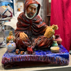 May include: A glazed ceramic figurine of a seated person wearing a dark red hooded robe and a white head covering. The figure holds a yellow vessel and is surrounded by several small, colorful ceramic pots on a textured blue base.