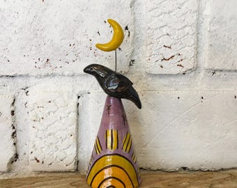 May include: A ceramic sculpture featuring a black bird perched atop a lavender cone-shaped base. The base is decorated with a yellow sun motif and small star accents. A yellow crescent moon is attached to a wire above the bird.