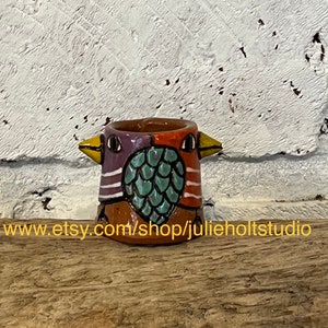 Pode incluir: Uma pequena xícara de cerâmica em forma de pássaro pintada à mão com um design turquesa, roxo, laranja e branco. A xícara tem uma pequena abertura no topo e um fundo arredondado.
