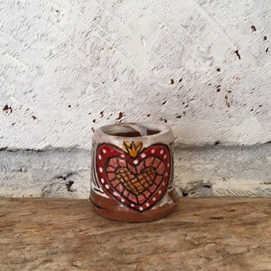 Könnte beinhalten: Eine kleine, braune Keramiktasse mit einem weißen Herzmotiv. Das Herz ist mit rosa und gelben Details und einer goldenen Krone verziert.