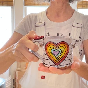 May include: A handmade ceramic mug with a rainbow heart design. The mug has a white base with black vertical lines and a red handle. The heart features concentric rings of various colors, including red, orange, yellow, green, blue, and purple.