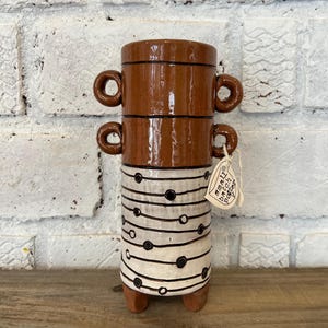 May include: A cylindrical ceramic vessel with two sets of loop handles on the sides. The top section is solid brown, while the bottom section is cream with horizontal black lines and dots. A tag reads small batch pottery. It stands on three feet.