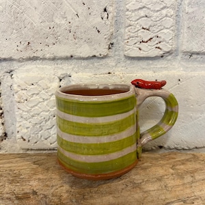 Peut inclure: Tasse en céramique faite à la main avec des rayures horizontales vertes et blanches. La poignée est rayée de rose et de blanc. Un petit poisson rouge en céramique est fixé sur le dessus de la poignée. La tasse est posée sur une surface en bois.