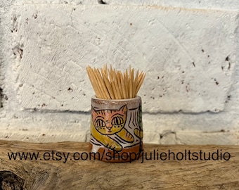 May include: Ceramic toothpick holder featuring a colourful cat design. The holder is filled with wooden toothpicks.