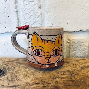 Peut inclure: Une tasse en céramique faite à la main avec un motif de visage de chat fantaisiste en orange et noir. La tasse a un fond gris avec des rayures noires et un accent de piment rouge sur la poignée. La tasse est posée sur une surface en bois.