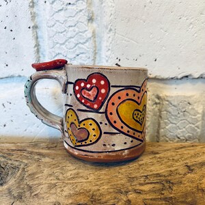 May include: Handmade ceramic mug with a light brown base and a gray body. The mug features colorful heart designs in red, yellow, and orange. The handle is a light blue and the top of the handle has a red accent.