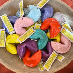 May include: A wooden bowl filled with colorful ceramic fortune cookies. The cookies are in various colors, including pink, yellow, blue, purple, and orange, with speckled finishes. Each cookie has a small tag that reads "MAY SUNSHINE & GOOD FORTUNE LIGHT YOUR WAY".
