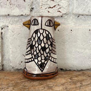 May include: A handmade ceramic vase with an owl design. The vase is primarily white with black and brown accents. The owl's face features large eyes and a beak, with a patterned body. The base is a reddish-brown color.