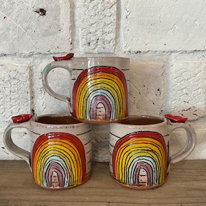Könnte beinhalten: Drei Keramikbecher mit Regenbogenmuster. Jeder Becher hat einen grauen Körper, einen gebogenen Henkel und einen roten Akzent am Rand. Das Regenbogenmuster weist leuchtende Farben auf, darunter Rot, Orange, Gelb, Grün, Blau und Violett.