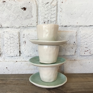 May include: A stack of three white ceramic cups and saucers. The cups have a textured, slightly uneven surface. The saucers are a light blue-green color.