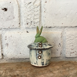 Op de afbeelding: Een keramische pot met een witte en blauwe glazuur en een groen keramiek konijnenbeeldje erop. De pot heeft een deksel met drie witte knoppen.
