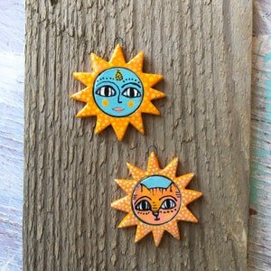 Puede incluir: Dos adornos de sol de cerámica con lunares naranja y amarillos. Un sol tiene una cara azul con un tercer ojo, el otro tiene una cara de gato naranja.