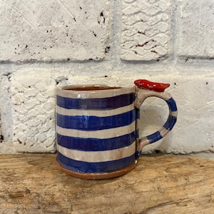 May include: A ceramic mug with blue and off-white horizontal stripes. The handle is striped blue and off-white, and a small red fish decorates the top of the handle. The mug sits on a wooden surface.