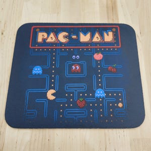 May include: A rectangular black mouse pad featuring a pixelated Pac-Man game screen. The design includes the orange Pac-Man logo, blue maze walls, yellow dots, a red ghost, two blue ghosts, a peach, cherries, and a strawberry on a dark background.