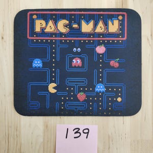 May include: A rectangular black mouse pad featuring a colorful pixelated Pac-Man game screen graphic. The design includes the yellow Pac-Man character, blue ghosts, a red ghost, fruit icons, and a blue maze layout with the text PAC-MAN at the top.
