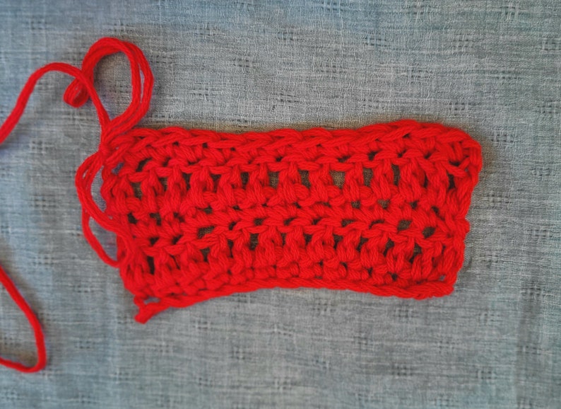 May include: A red crocheted rectangle with a loose end of yarn. The crochet is a simple stitch pattern.