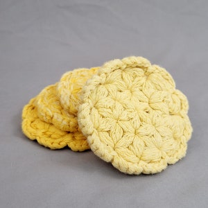 May include: A stack of four handmade, yellow crochet coasters. The coasters have a scalloped edge and a textured, floral-like pattern. The color is a soft, sunny yellow, and the coasters are arranged on a gray background.