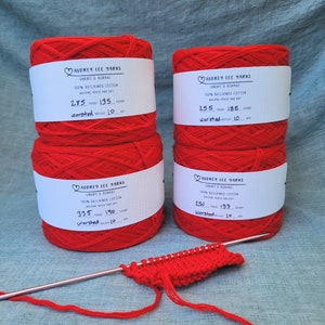 May include: Four spools of red yarn labeled "Audrey Lee Yarns" with details about the yarn, including the yarn weight, yardage, and grams. A knitting needle and a small swatch of red knit fabric are also pictured.